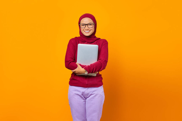 Portrait of smiling Asian woman bring laptop over yellow background - Photo, Image