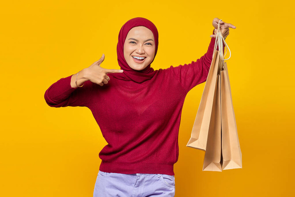 Beautiful Asian woman showing some shopping bags and giving thumbs up with smiling face on yellow background - Photo, Image