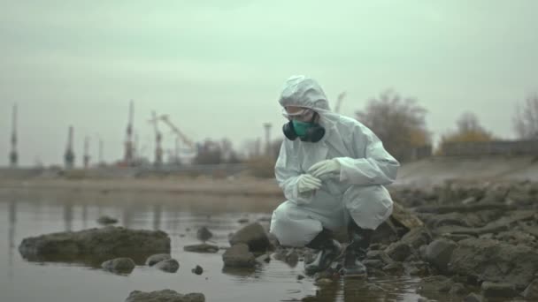 Slowmo πλάνο του θηλυκού οικολόγου με χημική στολή, αναπνευστική μάσκα, googles και γάντια που εξετάζουν την κατάσταση του νερού στην περιοχή βιολογικού κινδύνου, λαμβάνοντας δείγμα από αυτό σε δοκιμαστικό σωλήνα - Πλάνα, βίντεο