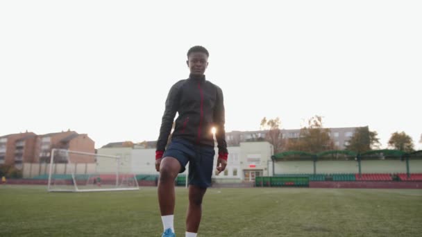 Jovem afro-americana de pé em um campo de futebol contra o fundo de um portão e colocando seu pé em uma bola de futebol - Filmagem, Vídeo