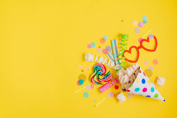 Holiday decor paper heart shaped eye glasses on stick for photosessions, lollipop and marshmallows with multicolored confetti near party hat on yellow background with blank space for text. Top view, flat lay. - Φωτογραφία, εικόνα