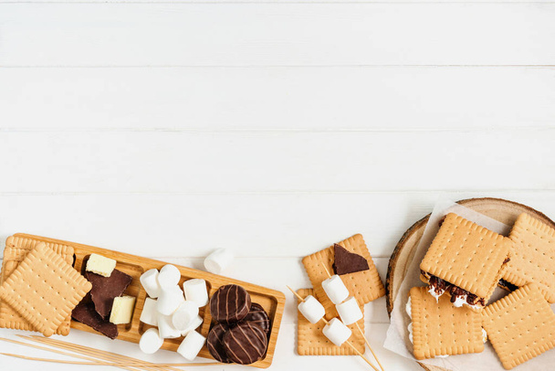 Marshmallow arrosto S'mores con cioccolato tra biscotti su sfondo bianco in legno con spazio vuoto per il testo. Vista dall'alto, piatta. - Foto, immagini