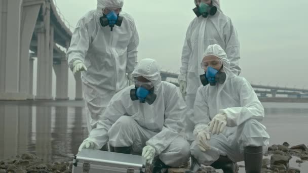Slowmo-Porträt eines multiethnischen Wissenschaftlerteams in Schutzanzügen, Atemschutzmasken und Brille, das auf verschmutzter Meeresküste sitzend in die Kamera blickt - Filmmaterial, Video