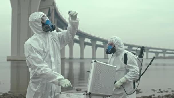 Slowmo shot of African-American male scientist in chemical suit looking at test tube with polluted soil sample and his colleagues exploring seashore of biohazard territory - Footage, Video