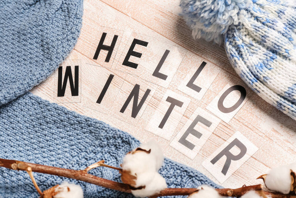Composición con texto HELLO WINTER, sombrero cálido, suéter y rama de algodón sobre fondo de madera clara, primer plano - Foto, imagen