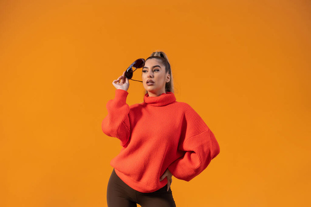 A trendy girl in a bright orange sweater and black sunglasses posing in the bright background - Foto, Imagem