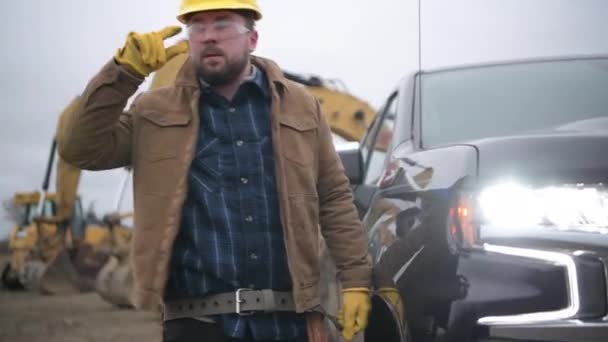 Empreiteiro de Construção Caucasiano Trabalhador em seus 30 anos com cinto de ferramentas ficar ao lado de seu caminhão. Escavadeiras em segundo plano. - Filmagem, Vídeo