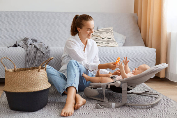 Full length portrait of positive happy female with ponytail wearing white shirt and jeans playing with her toddler daughter or son while sitting on floor near gray sofa. - Fotografie, Obrázek