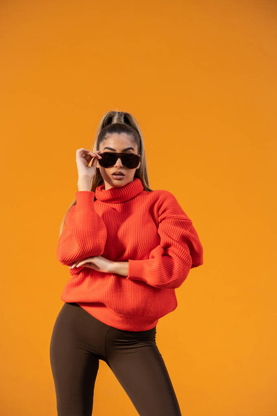 A sexy female in a trendy outfit and black sunglasses is posing in the orange background - Fotó, kép