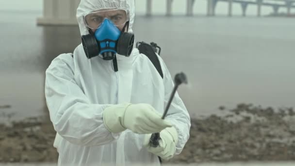 湖の岸の領土上に特別な液体を噴霧化学的な防虫スーツ、呼吸マスクやグーグルで白人男性科学者の中低速ショット - 映像、動画
