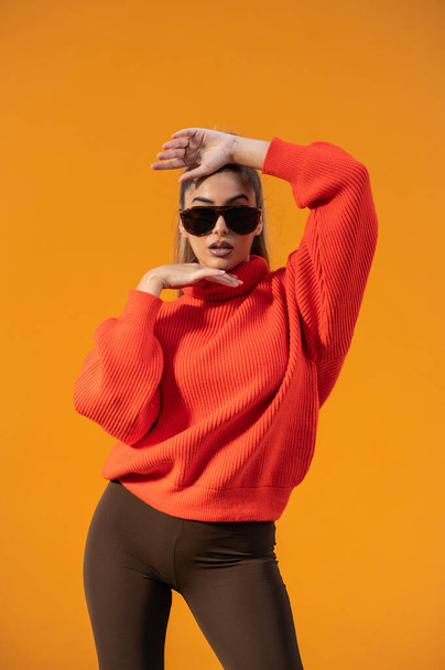 An attractive slim model in a trendy outfit and black sunglasses is posing in the orange background - Fotoğraf, Görsel