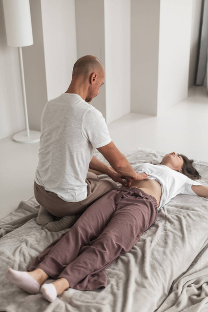 A professional young man massaging a woman's stomac in a white cozy room during a manual therapy - Foto, Imagem