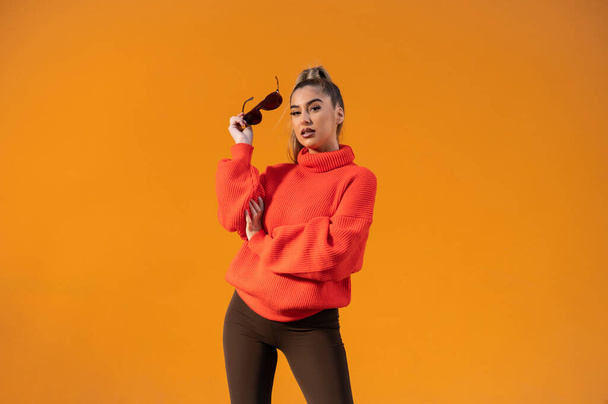 A beautiful Spanish lady wearing an orange sweater and holding blasunglasses is posing in the orange background - Фото, зображення