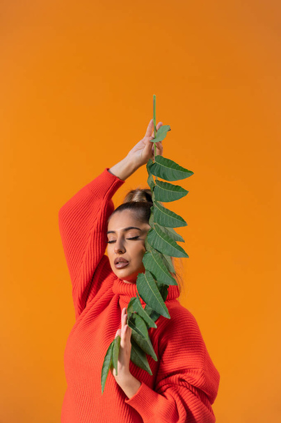 A vertical shot of a Spanish female with eyes-closed posing with a plant branch in the bright orange background - Foto, immagini