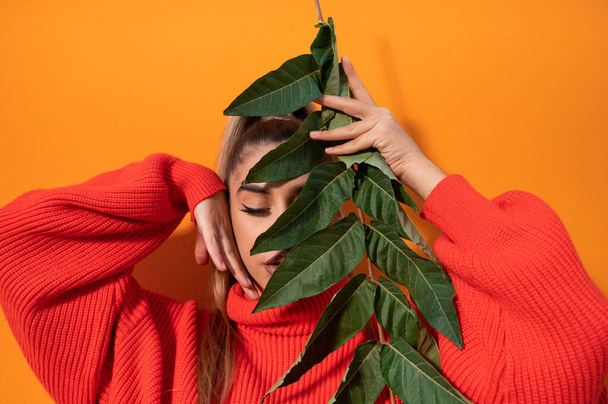 A stylish young female with eyes-closed is holding a branch with green leaves in the orange background - Foto, afbeelding
