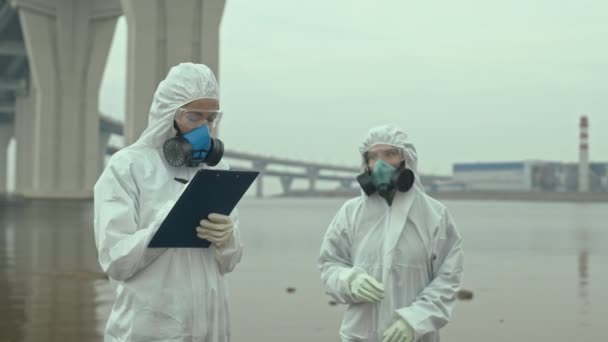 Slowmo shot of two female ecology experts in protective coveralls discussing water sample one holding in hand standing outdoors on biohazard territory - Footage, Video