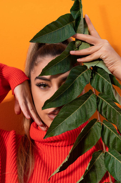 A vertical shot of a Spanish girl wearing an orange sweater and holding a branch with green leaves - Foto, afbeelding