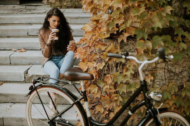 Pretty young woman with mobile phone drink takeaway coffee at the stairs by the bicycle on autumn day - Photo, Image