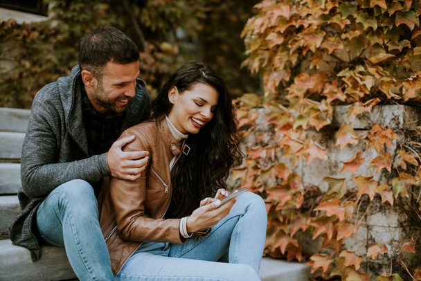Handsome young couple sitting on outdoor stairs on a autumn day and using mobile phone - Photo, Image