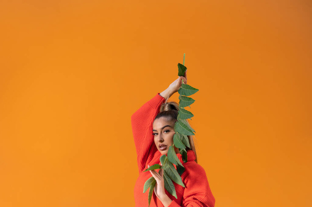 A stylish Spanish girl in a casual orange sweater is holding a tree branch with leaves; orange background with a copy space - Фото, изображение