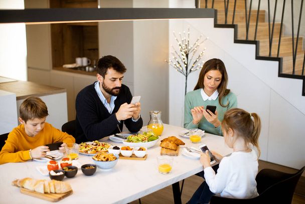 Famille utilisant des téléphones portables tout en prenant le petit déjeuner à la table à manger à l'appartement - Photo, image