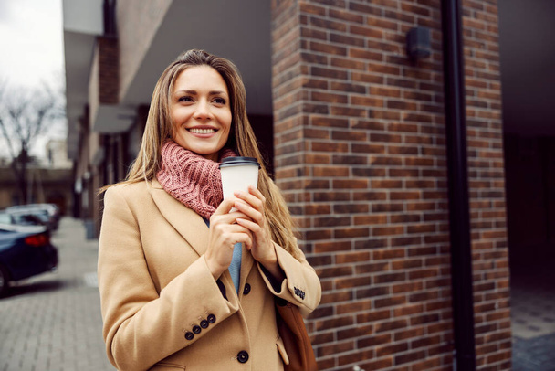 Μια χαρούμενη νεαρή γυναίκα με παλτό και κασκόλ κρατάει ζεστό καφέ στα χέρια της. Τις κρύες μέρες, έχουμε ένα ζεστό ρόφημα να μας ζεστάνει. Μια γυναίκα με καφέ απ 'έξω.. - Φωτογραφία, εικόνα