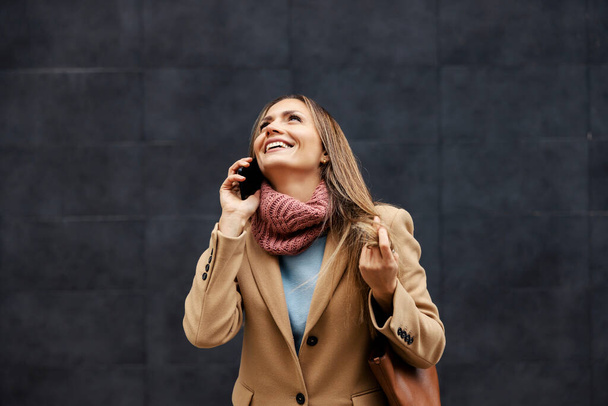 A happy young fashionable woman is standing outdoors in a warm outfit and having a phone conversation. Global communications. A woman talking on the phone. - Foto, Bild