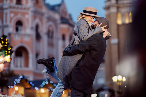 Amantes de la Navidad abrazándose en la calle en Año Nuevo. Un hombre feliz está de pie en la calle, levantando a su novia y abrazándola en Nochebuena. - Foto, imagen