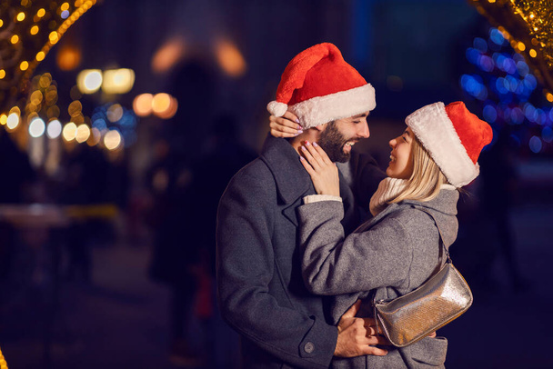 Різдвяна пара обіймає на свіжому повітрі напередодні Різдва. Молода щаслива пара закохана в Санта-Капелюхи на голові стоїть на вулиці напередодні Різдва і обіймає
. - Фото, зображення
