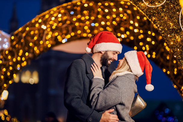 Weihnachtsliebhaber umarmen und lachen sich an Heiligabend auf der Straße. Romantische Momente und festliche Stimmung. Ein junges glückliches Paar mit Nikolausmützen auf dem Kopf umarmt sich an Heiligabend. - Foto, Bild