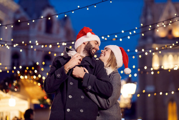 Karácsonyi szerelmesek nevetnek és ölelkeznek az utcán karácsony este. Egy fiatal boldog pár Télapó sapkával a fején áll az utcán, és karácsony este ölelkeznek.. - Fotó, kép