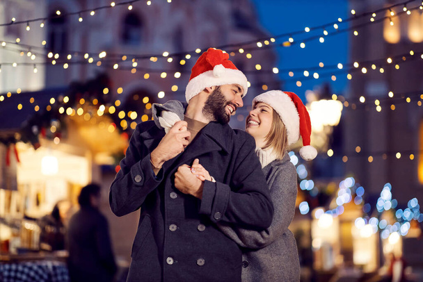 Christmas spouses hugging on the street on New Year. A young happy couple in love with Santa hats on their heads is standing on the street, hugging and looking at each other on New Year's eve. - Photo, Image