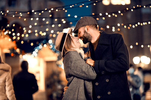 Kerstmis echtgenoten kussen en knuffelen op straat op kerstavond. Een jong gelukkig stel staat 's nachts op straat, knuffelend en kussend op kerstavond. - Foto, afbeelding