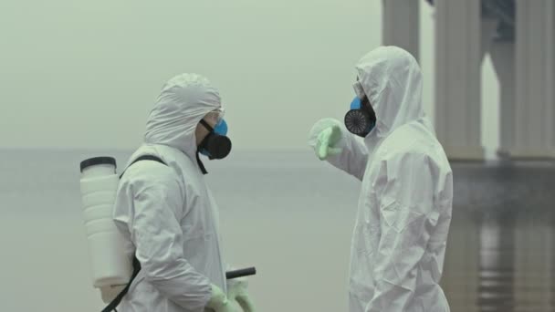 Medium slowmo portrait of two diverse scientists in chemical suits and respiratory masks discussing elimination of biohazard standing in zone where eco catastrophe has happened then looking at camera - Footage, Video