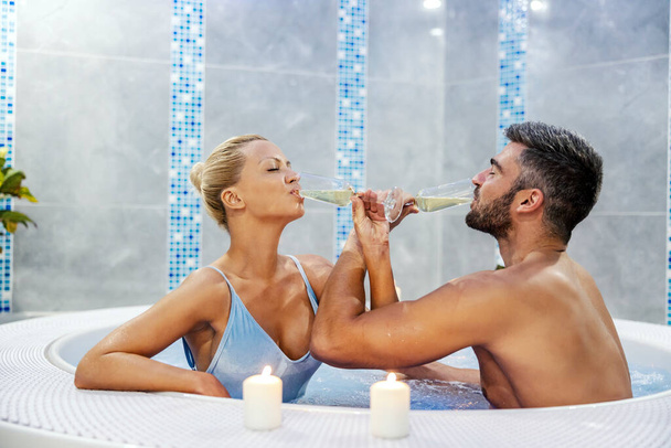 A romantic couple sitting in the hot tub at the spa center and drinking champagne with arms crossed. Moments together at weekend. A couple drinking champagne in the hot tub. - Фото, изображение