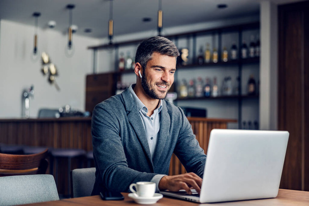 スマートカジュアルを着たビジネスマンは、耳の中のイヤホンでカフェに座って、ノートパソコンにレポートを入力しています。コーヒーショップでノートパソコンを使う現代のビジネスマン. - 写真・画像