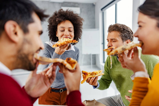 Biznesmeni jedzą lunch w biurze. Mała grupa wielorasowych kolegów siedzących w biurze i delektujących się pizzą do płuca podczas przerwy obiadowej. - Zdjęcie, obraz