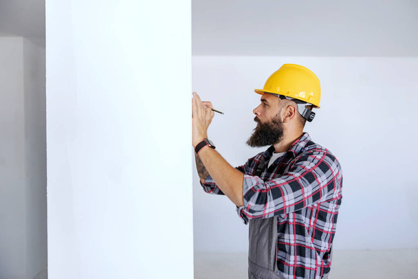 A worker in overalls and helmet on his head is standing and writing on the wall where to drill a hole for electric installations. - Fotoğraf, Görsel