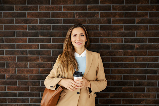 A happy woman with long hair in a warm outfit leaning on the brick wall outside and holding her fresh morning takeaway coffee. The morning routine of a woman. A woman holding takeaway coffee. - Photo, Image
