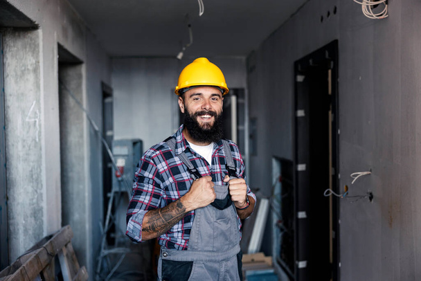 A happy, hardworking handyman with a helmet on his head holds his overalls and looking at the camera. There is a lot of work to do, but he is inspired and happy to get to work. - Фото, изображение