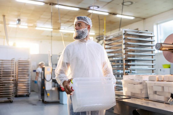 Un operaio di una fabbrica alimentare in uniforme bianca sterile sta trasferendo contenitori di plastica. Se il prodotto della fabbrica è alimento, dobbiamo essere sicuri che tutto sia sterile e pulito alla perfezione. - Foto, immagini
