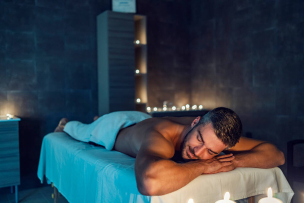 Un hombre relajado disfruta momentos después del masaje en el centro de spa. A su alrededor hay velas fragantes. Aromaterapia y masaje. Un hombre en el masaje en el centro de spa. - Foto, imagen