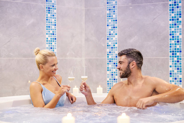 Una feliz pareja está sentada en la bañera de hidromasaje del centro de spa y brindando con champán. La pareja está rodeada de velas. Fin de semana romántico en el resort. Pareja en el jacuzzi - Foto, Imagen