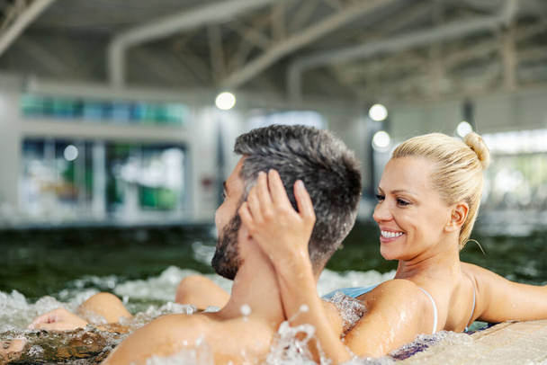 Une femme blonde souriante assise avec son petit ami bien-aimé dans la piscine avec de l'eau thermale au centre de spa et le câlinant. Une femme heureuse qui montre de l'amour. Spa et amour. Couple au centre de spa dans la piscine. - Photo, image