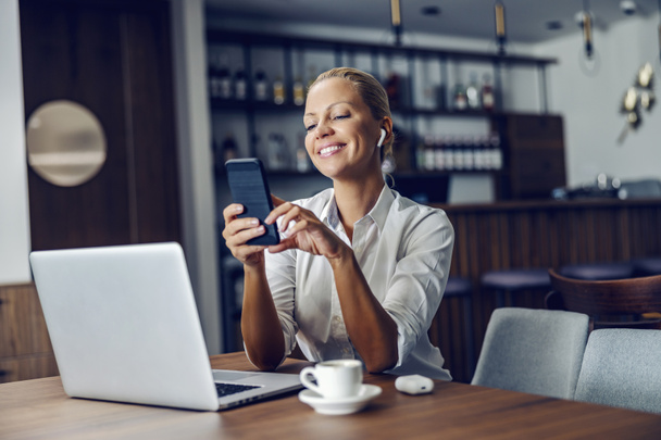A happy elegant businesswoman with earphones sits in a cafe and using her phone for a video call. She is taking a break from work. There is a laptop on the table. A businesswoman having a video call - Foto, Imagem