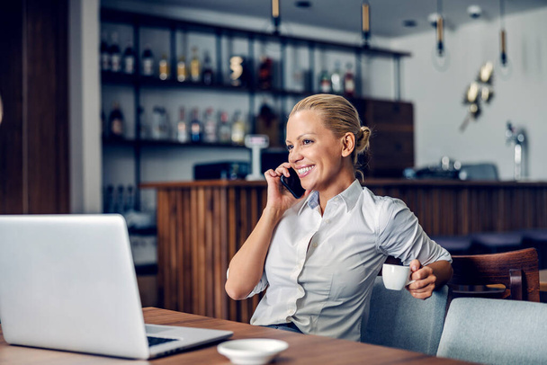Μια ευτυχισμένη επιχειρηματίας με επίσημα ρούχα κάθεται σε ένα φιλικό καφέ εργασίας και έχει μια σημαντική κλήση. Λύνει ένα πρόβλημα με ελάχιστη προσπάθεια. Η γυναίκα κρατάει καφέ. Μια επιχειρηματίας που εργάζεται σε ένα καφέ - Φωτογραφία, εικόνα