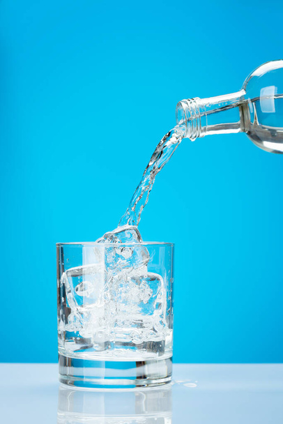 Свежая вода наливается из бутылки в стекло с кубиками льда на синем фоне - Фото, изображение