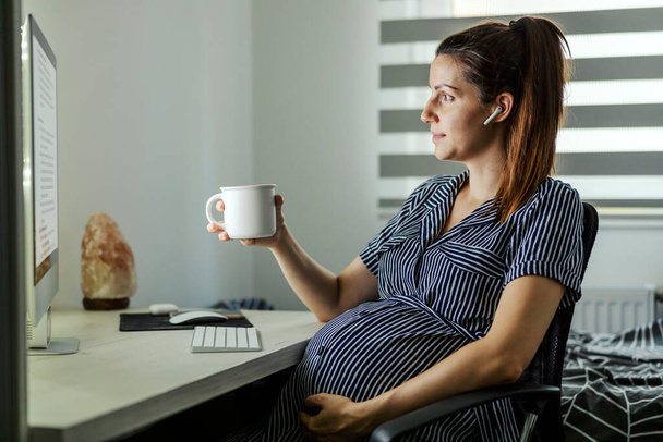 Вагітна жінка п'є гарячий напій в домашньому офісі. Успішна вагітна жінка з навушниками в смугастій сукні сидить перед комп'ютером з навушниками в вухах і використовує телефон
 - Фото, зображення