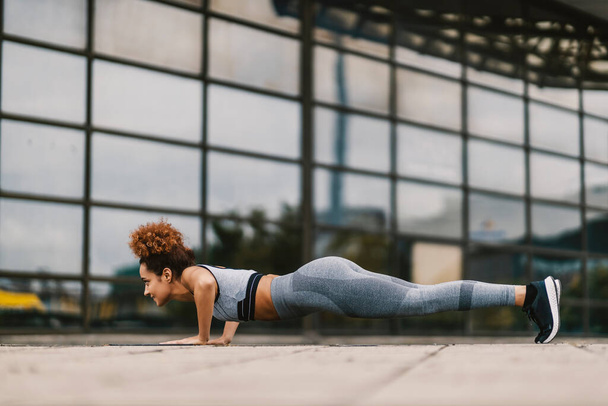 Erős, sportos nő fekvőtámaszt csinál a város üvegépülete előtt. Egy városi sportoló fekvőtámaszt csinál.. - Fotó, kép