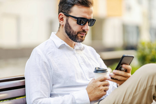 Porträt eines bärtigen Geschäftsmannes mittleren Alters mit Sonnenbrille, der auf einer Bank sitzt und auf seinem Handy blättert. Er macht eine Kaffeepause, also trinkt er Kaffee to go. - Foto, Bild